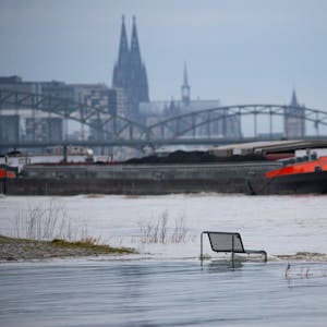 Binnenschiffe fahren auf dem Rhein vor der Kulisse des Kölner Doms, im Vordergrund steht eine vom Wasser umspülte Sitzbank.