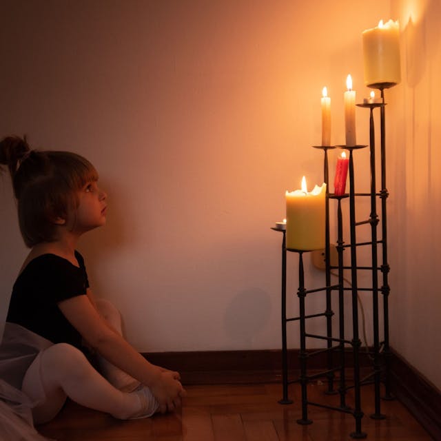 Ein kleines Mädchen sitzt im Schneidersitz auf dem Boden und betrachtet einen Kerzenständer mit brennenden Kerzen.&nbsp;