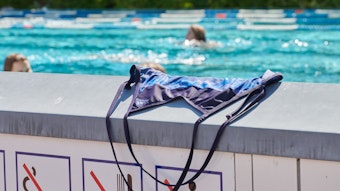 Ein Badeanzug liegt auf einer Mauer in einem Schwimmbad, dahinter schwimmen Menschen im Becken ihre Bahnen.