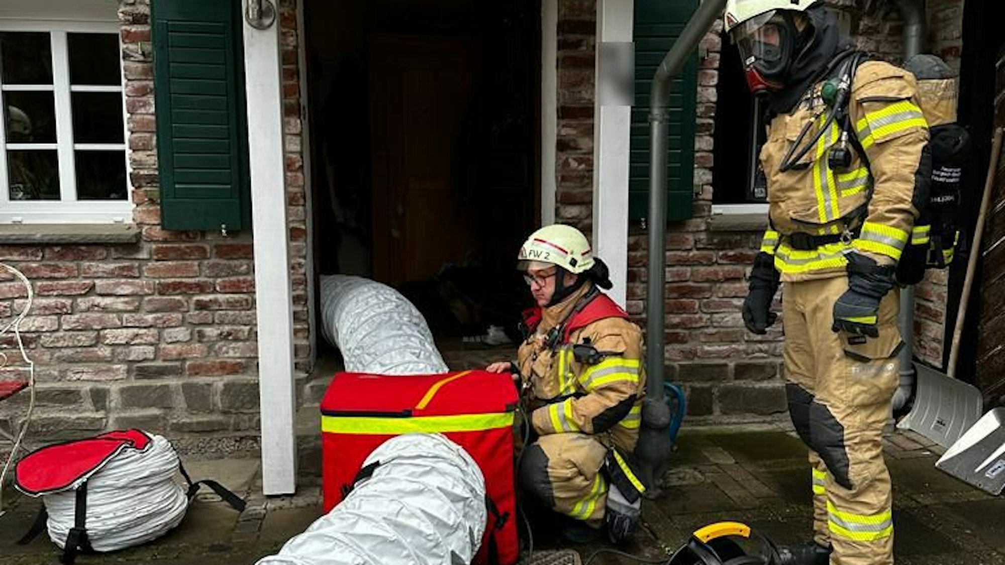 Zwei Feuerwehrleute in Schutzausrüstung installieren ein großes Lüftungsgebiet mit Schläuchen, die in ein Haus hineinführen.