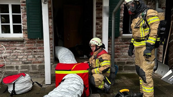 Zwei Feuerwehrleute in Schutzausrüstung installieren ein großes Lüftungsgebiet mit Schläuchen, die in ein Haus hineinführen.