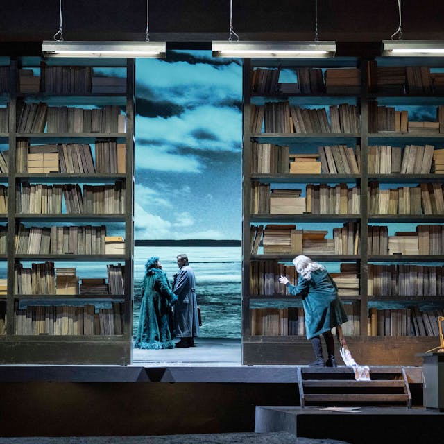 Auf der Bühne stehen zwei sehr hohe Bücherschränke, in der Mitte zeigt ein Spalt einen Strand, wo ein Paar sich an den Händen hält.
