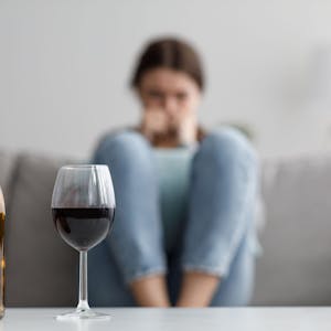 Im Hintergrund sitzt eine Frau auf einem Sofa, davor stehen eine leere Flasche Wein und ein volles Wein-Glas auf dem Tisch