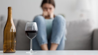 Im Hintergrund sitzt eine Frau auf einem Sofa, davor stehen eine leere Flasche Wein und ein volles Wein-Glas auf dem Tisch
