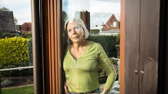 Eine Frau steht in ihren Haus und blickt durch die Fensterscheibe in ihren Garten.