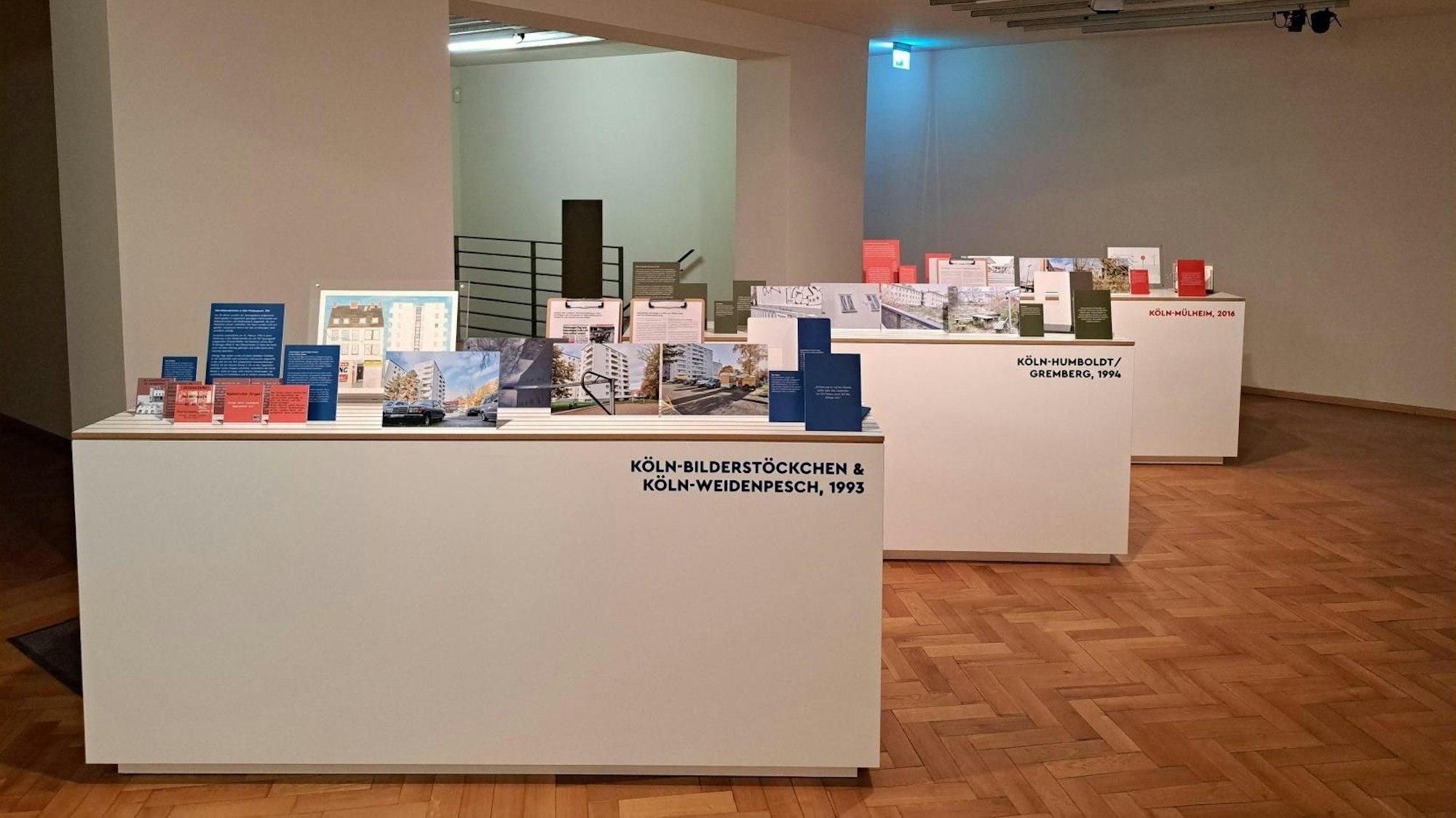 Ansicht auf einen Nebenraum der Ausstellung. Auf drei einzelnen länglichen Tischen sind jeweils Fotografien, Texte und Informationen zu Orten rechter Gewalt in Köln aufgestellt.