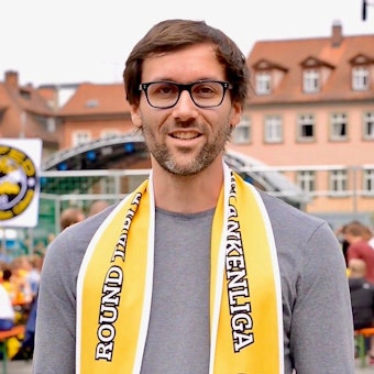 Der Vorstand des „Team Bananenflanke Köln e.V.“: Maximillian Küsters ist im Porträt zu sehen. Er trägt einen Dreitagebart, eine Brille und hat kurzes braunes Haar. Er lächelt in die Kamera.