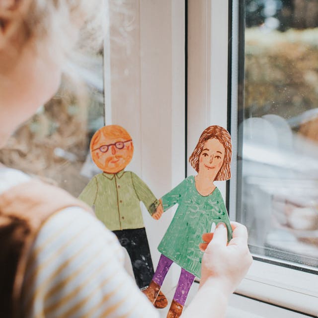 Ein kleines Mädchen steht am Fenster und hält zwei bemalte Pappfiguren in der Hand, eine Frau und einen Mann mit Bart und Brille.&nbsp;