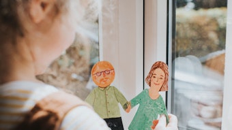 Ein kleines Mädchen steht am Fenster und hält zwei bemalte Pappfiguren in der Hand, eine Frau und einen Mann mit Bart und Brille.