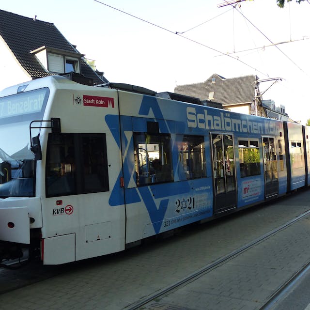 Durch eine Frechener Straße fährt eine KVB-Straßenbahn der Linie 7 mit blau-weißem Schriftzug "Schalömchen Köln".