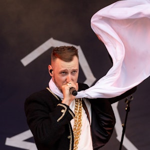 Der deutsche Rapper Alligatoah steht beim Open-Air-Festival "Rock im Park" auf der Bühne. Er singt in ein Mikrofon. Er trägt einen weißen Schal, der im Wind weht.