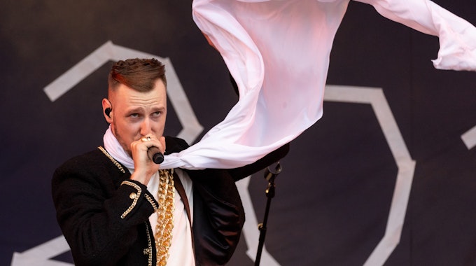 Der deutsche Rapper Alligatoah steht beim Open-Air-Festival "Rock im Park" auf der Bühne. Er singt in ein Mikrofon. Er trägt einen weißen Schal, der im Wind weht.
