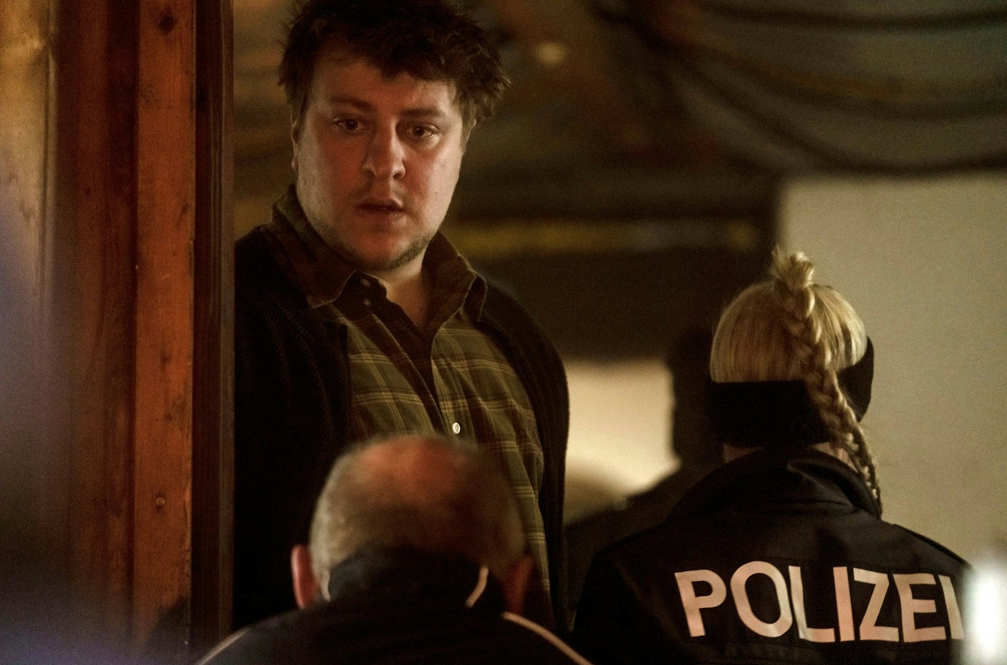 Matthias Precht schaut verdutzt, während Polizeibeamte an ihm vorbei in sein aus gehen. Der erzieher trägt ein mattgrünes, gestreiftes Hemd.