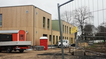 An einem Bauzaun vorbei ist ein zweistöckiges Gebäude mit einer Front aus Holz zu sehen. Die Kindertagesstätte soll im Sommer eröffnet werden.