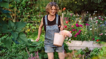 Anja Klein trägt eine Jeans-Latzhose und ein schwarzes T-Shirt und hält einen rosafarbenen Eimer mit Blumen in der Hand.