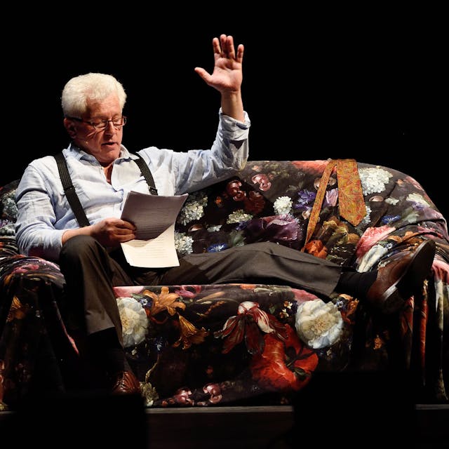 Schauspieler Miroslav Nemec sitzt auf einem Sessel auf der Bühne. Er hat ein Bein hochgelegt und liest von Zetteln. Mit der linken Hand gestikuliert er.
