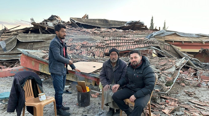 Der Euskirchener Ali Demirbas und zwei weitere Männer sitzen auf Plastikstühlen, um sie herum die Trümmer zerstörter Häuser.
