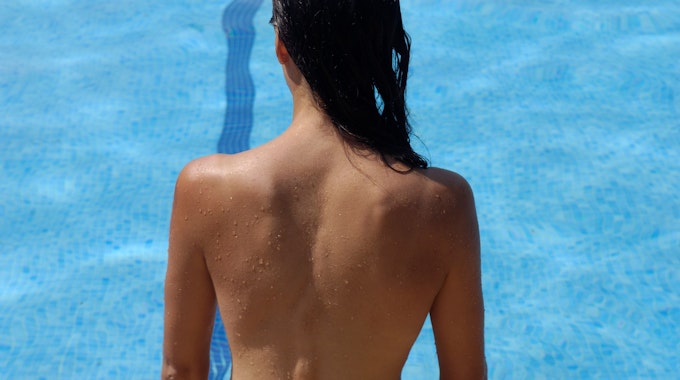 Eine Frau ist von hinten zu sehen. Sie steht ohne Bikini-Oberteil vor einem Schwimmbecken