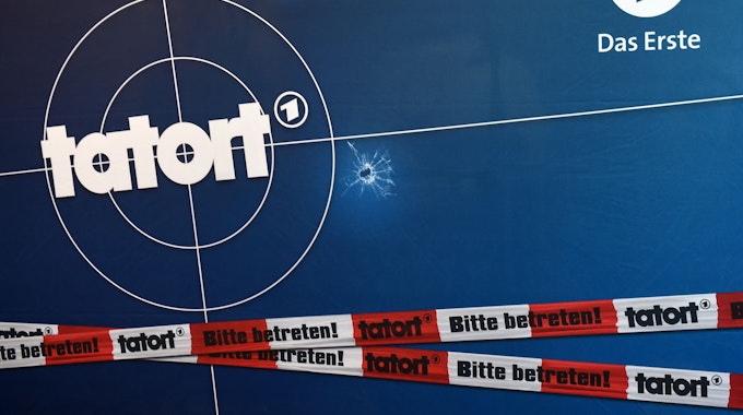 Auf einer Fotorückwand sind das Logo der ARD-Fernsehreihe „Tatort“, Einschusslöcher und Absperrband abgebildet.