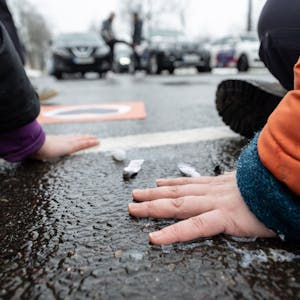 Klimaschutzaktivisten der „Letzten Generation“ kleben sich mit Sekunden kleben auf dem Asphalt einer Straße fest. Im Hintergrund stehen Autofahrer deswegen im Stau.