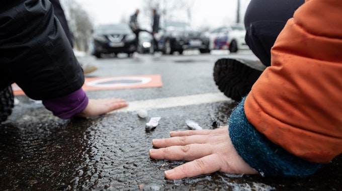 Klimaschutzaktivisten der „Letzten Generation“ kleben sich mit Sekunden kleben auf dem Asphalt einer Straße fest. Im Hintergrund stehen Autofahrer deswegen im Stau.