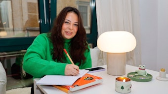 Cibelle Lopes  (25) trägt einen grünen Pullover und schreibt etwas in ein Heft, das auf einem Tisch vor ihr liegt.