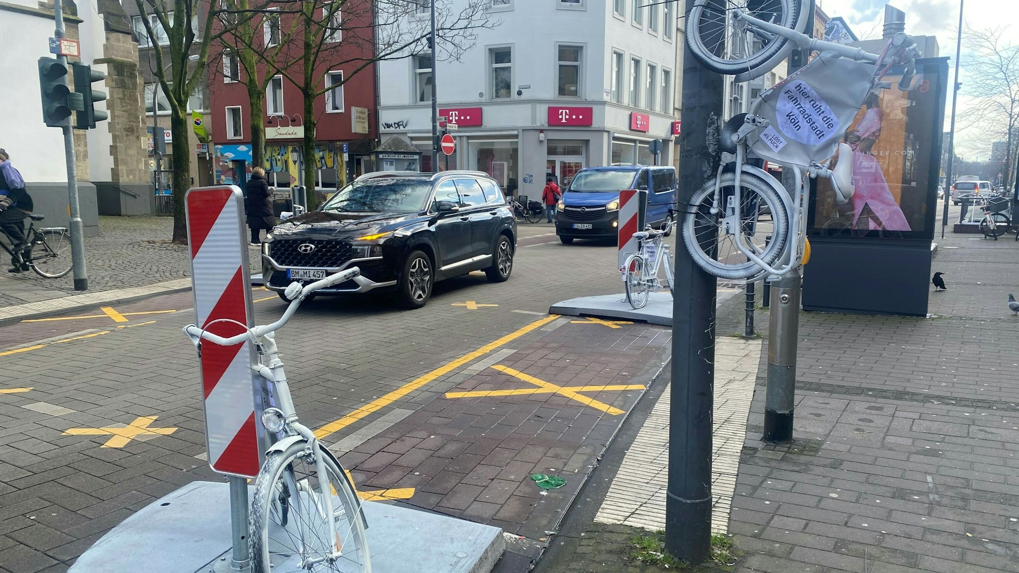 Auf der Venloer Straße stehen zwei weiße Fahrräder am Straßenrand, ein drittes hängt an einem Laternenmast.