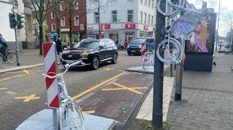Auf der Venloer Straße stehen zwei weiße Fahrräder am Straßenrand, ein drittes hängt an einem Laternenmast.
