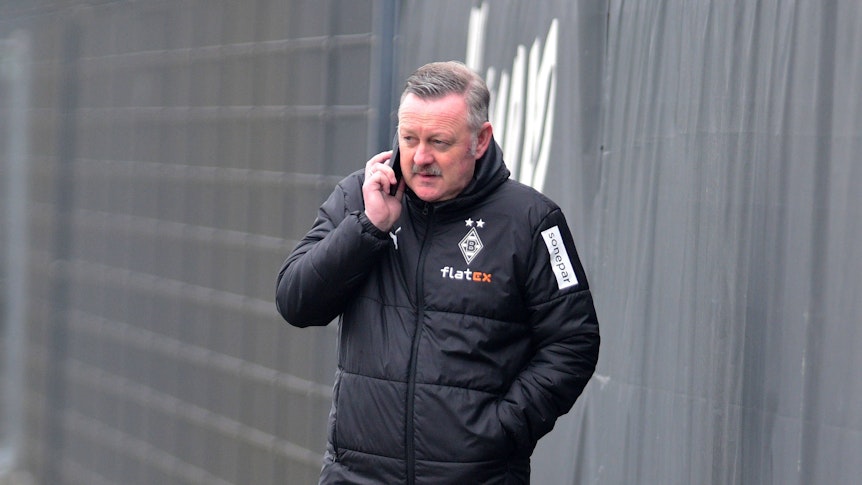 In Gesprächen: Borussia-Manager Roland Virkus telefoniert auf dem Trainingsplatz von Borussia Mönchengladbach während einer Einheit am 9. März 2023. Das Foto zeigt Virkus in einer Gladbach-Winterjacke und mit einem Telefon am rechten Ohr.