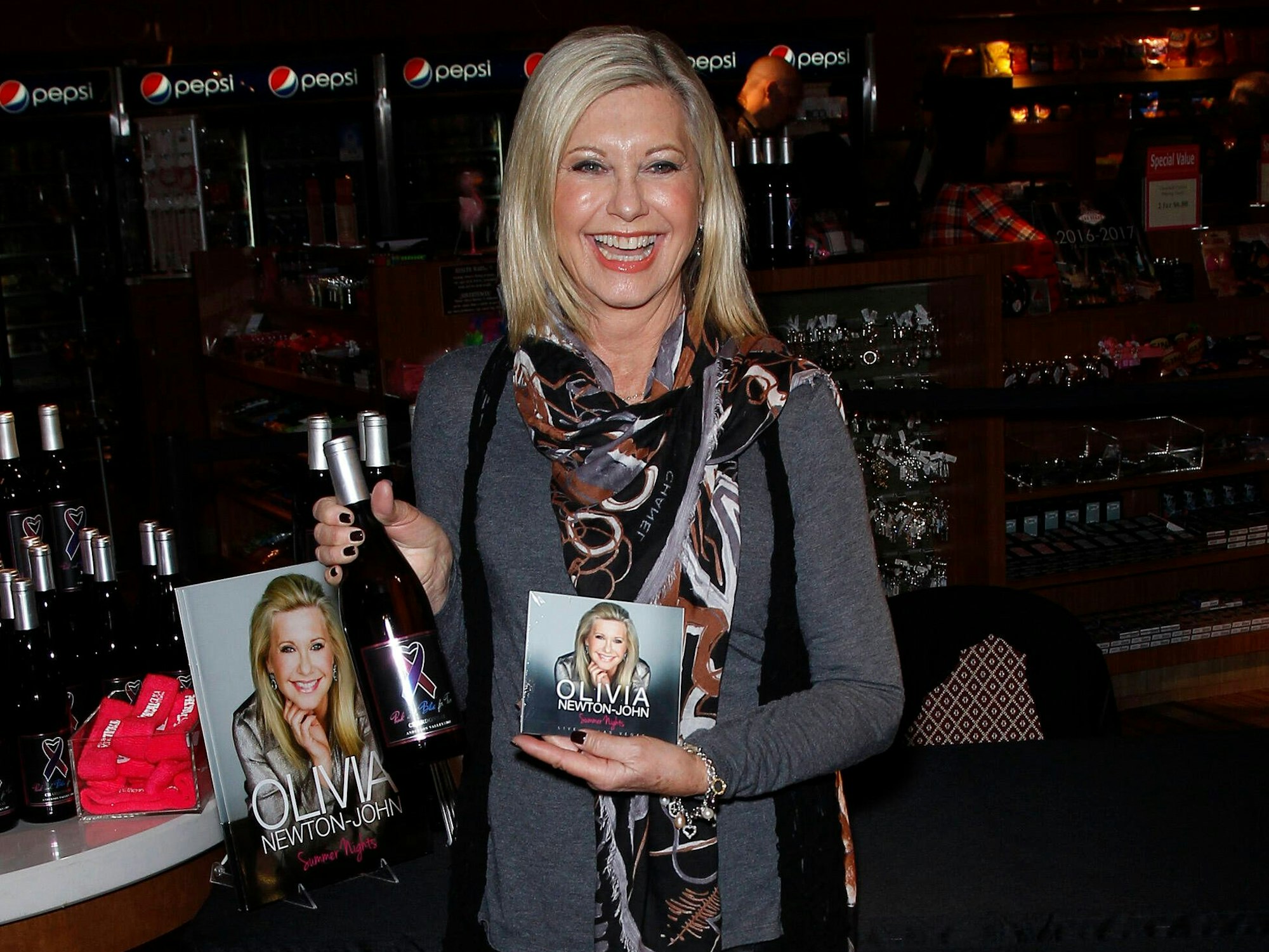 Olivia Newton-John hält ein Album in der linken und ihren Wein in der rechten Hand. Im Hintergrund sieht man auch ihr Buch und weitere Weinflaschen.