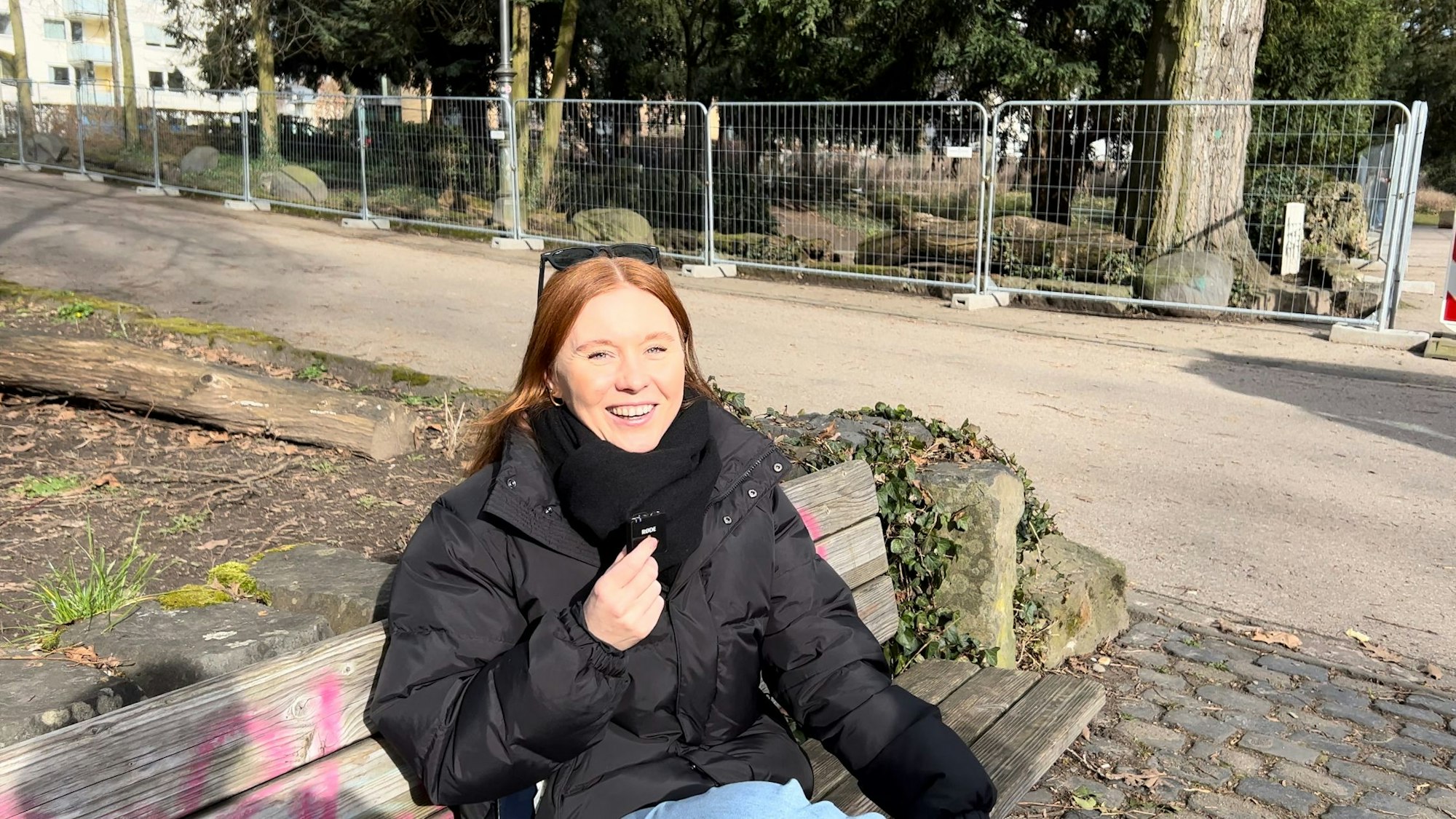 Eine Frau mit schwarzer Jacke sitzt auf einer Parkbank.