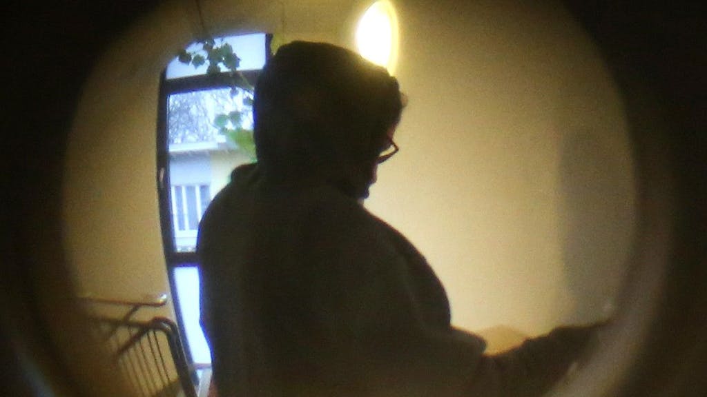 Durch einen Türspion ist eine Person mit einem Kapuzenpulli vor einer Wohnungstür zu sehen. Die Kölner Polizei sucht einen Mann, der möglicherweise am Tat-Tag einen schwarzen Kapuzenpullover trug. Das Foto steht in keinem Zusammenhang mit den aktuellen Vorkommnissen in Leverkusen.