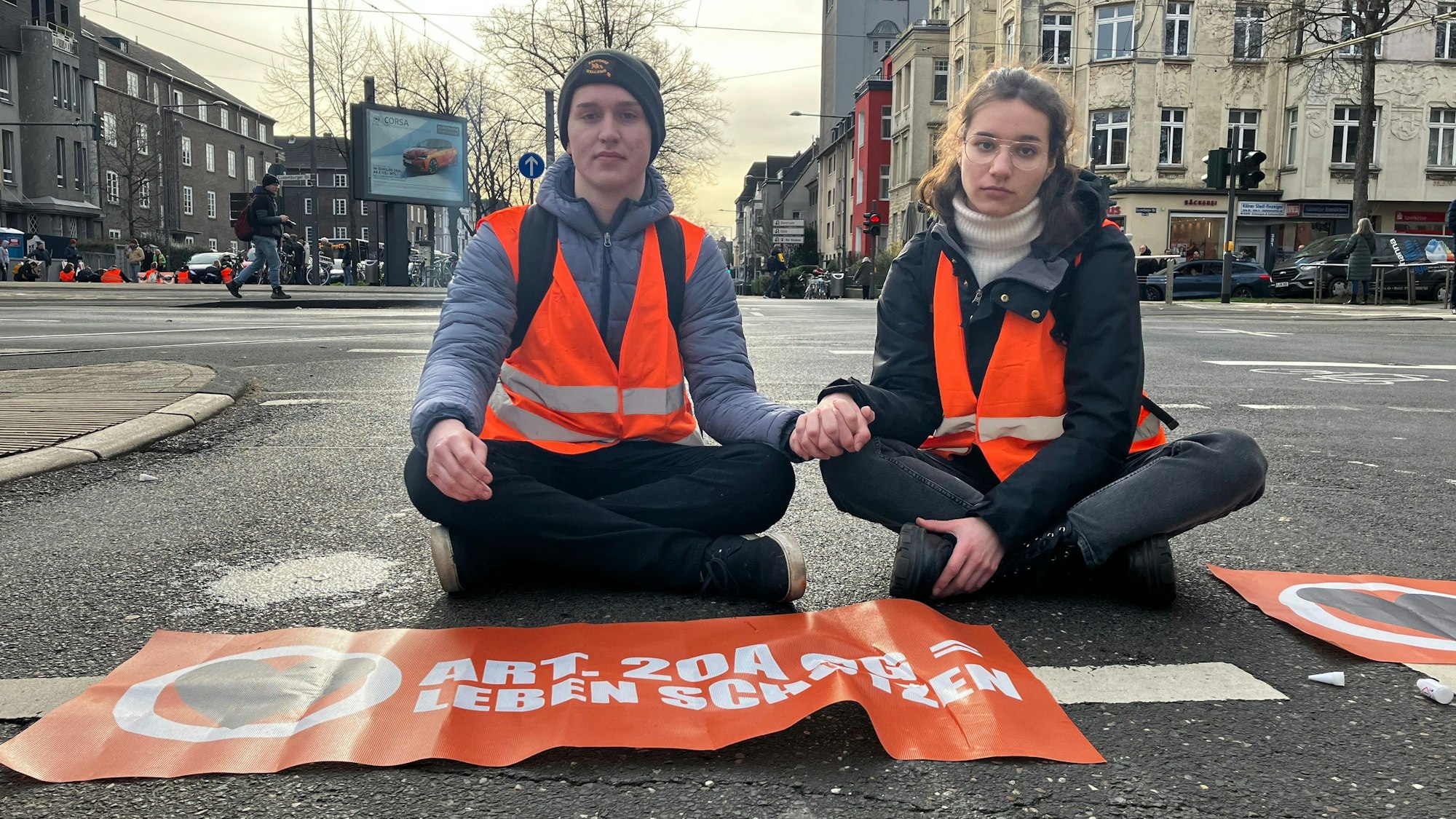 DIe Protestierenden sitzen versetzt auf den Straßen. Ein Aktivist und eine Aktivistin sitzen Hand in Hand auf der Straße.
