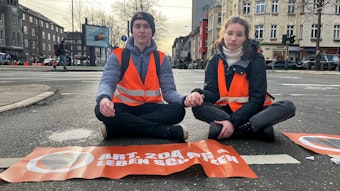 DIe Protestierenden sitzen versetzt auf den Straßen. Ein Aktivist und eine Aktivistin sitzen Hand in Hand auf der Straße.