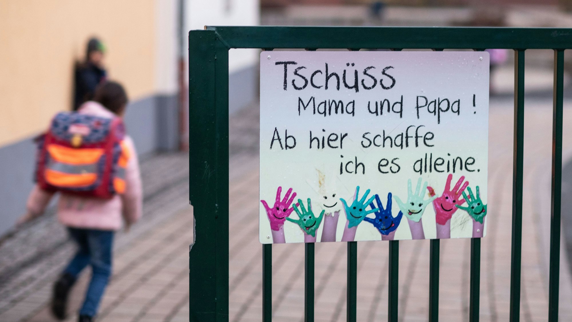Grundschüler gehen morgens auf den Schulhof, vorbei an einem Schild mit der Aufschrift „Tschüss Mama und Papa! Ab hier schaffe ich es alleine.“
