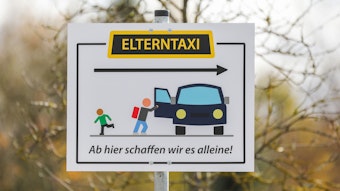 Ein Schild mit der Aufschrift „Elterntaxi Ab hier schaffen wir es alleine!“ steht am Parkplatz eines Schulgeländes.