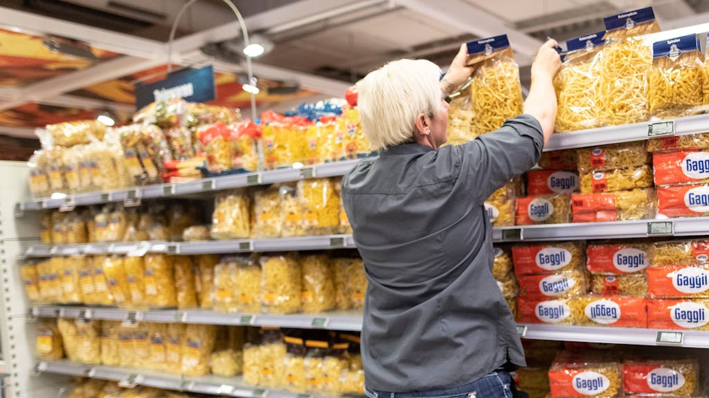 Das Symbolfoto aus dem Jahr 2020 zeigt eine Supermarkt-Mitarbeiterin mit grauem Hemd und kurzen blonden Haaren vor einem Regal mit allen möglichen Nudelsorten.