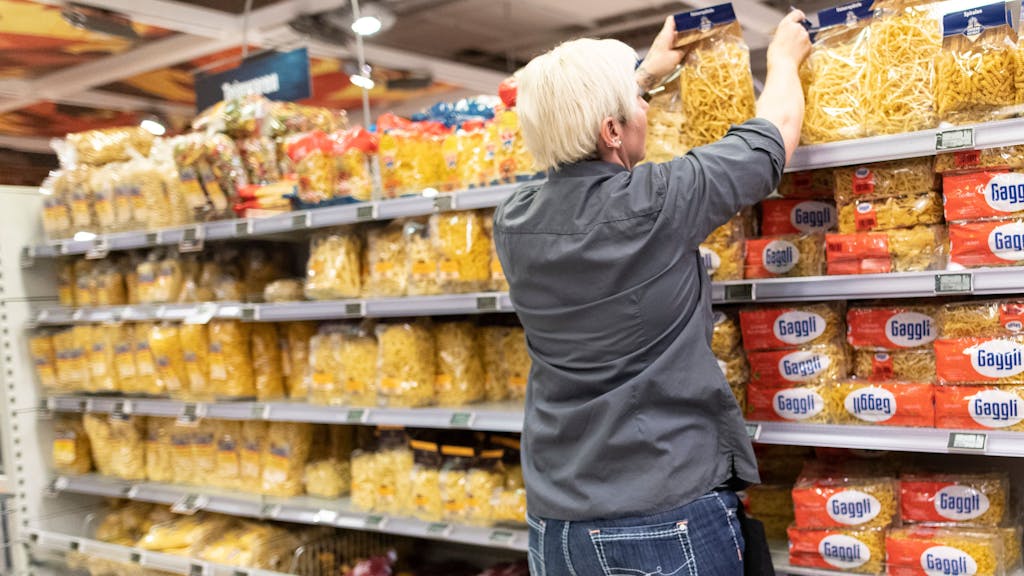 Das Symbolfoto aus dem Jahr 2020 zeigt eine Supermarkt-Mitarbeiterin mit grauem Hemd und kurzen blonden Haaren vor einem Regal mit allen möglichen Nudelsorten.