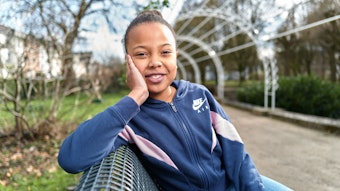 Sophia sitzt auf einer sonnigen Bank im Park und lächelt in die Kamera. Sie stützt ihren Kopf auf ihrer Hand auf.