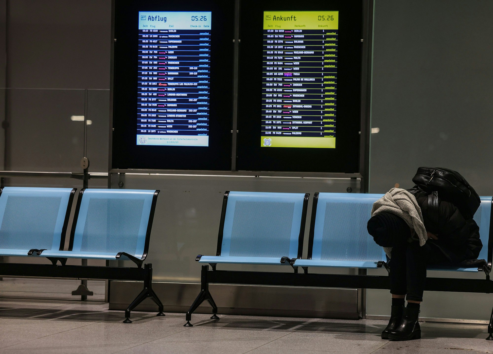 Eine Frau sitzt am Flughafen Köln/Bonn neben einer Anzeigetafel, auf der zahlreiche Flüge annulliert sind.