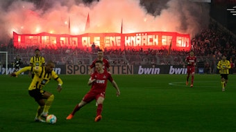 Spieler des FC und des BVB während des Spiels, im Hintergrund haben Fans des FC Leuchtfackeln angezündet und halten ein Banner hoch.