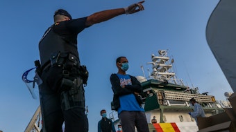 Flüchtlinge verlassen ein Schiff. Ein Mann zeigt ihnen die Richtung, in die sie gehen sollen.