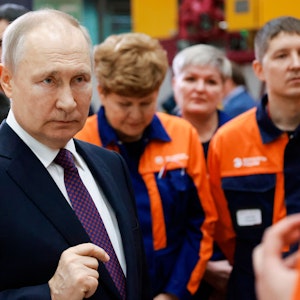 Das Foto aus dem Jahr 2023 zeigt den russischen Präsidenten Wladimir Putin. Er trägt einen dunklen Anzug, hinter ihm stehen mehrere Menschen in orange-blauen Uniformen.