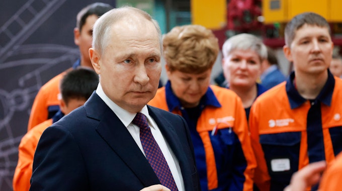 Das Foto aus dem Jahr 2023 zeigt den russischen Präsidenten Wladimir Putin. Er trägt einen dunklen Anzug, hinter ihm stehen mehrere Menschen in orange-blauen Uniformen.