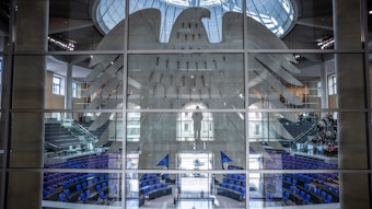Blick durch Glasscheiben in den leeren Plenarsaal im Bundestag. Unmittelbar hinter der Fensterfront prangt der riesige Bundesadler, im Bildhintergrund sind leere Sitzreihen des Plenarsaals sowie darüber Besuchersitzreihen zu sehen.