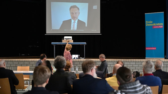 Menschen sitzen in einem Saal und schauen auf eine Leinwand, auf der im Videobild FDP-Bundesparteichef und Finanzminister Christian Lindner zu sehen ist.