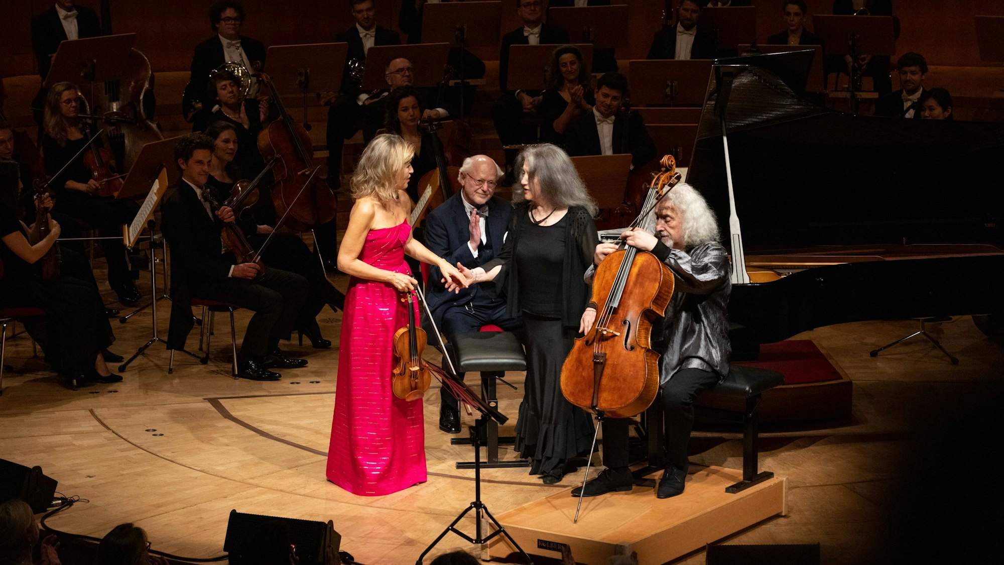 Anne-Sophie Mutter trägt ein magentafarbenes Kleid und gibt Martha Argerich die Hand. Daneben sitzt Mischa Maisky mit seinem Cello.
