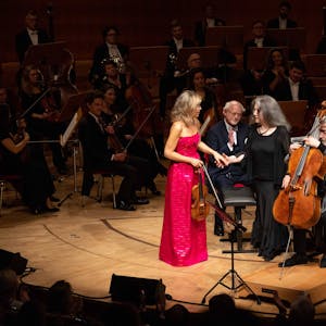 Anne-Sophie Mutter trägt ein magentafarbenes Kleid und gibt Martha Argerich die Hand. Daneben sitzt Mischa Maisky mit seinem Cello.&nbsp;