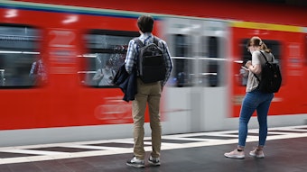 Zwei Studierende stehen am Bahnsteig, wo gerade ein Zug durchfährt.