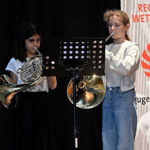 Die beiden stehen auf einer Bühne mit ihren Instrumenten, links im Bild ein Banner.